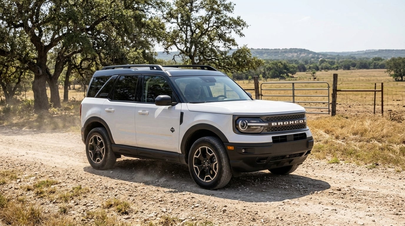 A white Ford Bronco Sport SUV driving along a dusty dirt road past a farm gate and trees on a sunny day. (Representative image for illustration purposes only. Actual vehicle may vary based on trim and configuration.)